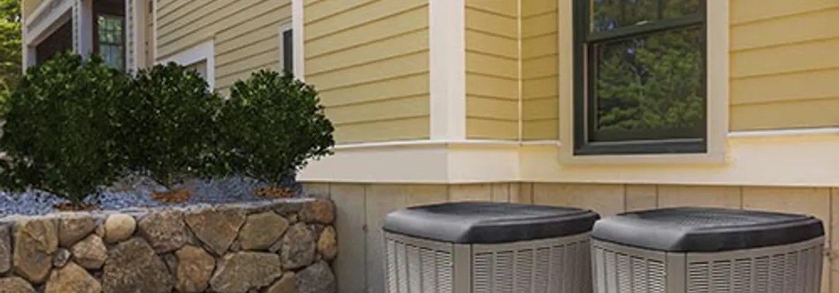 Two outdoor heat pump units on a gravel bed beside a timber-clad house